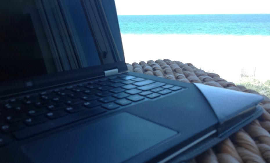 laptop sitting on a wicker table overlooking an ocean of emarld and sand of white.